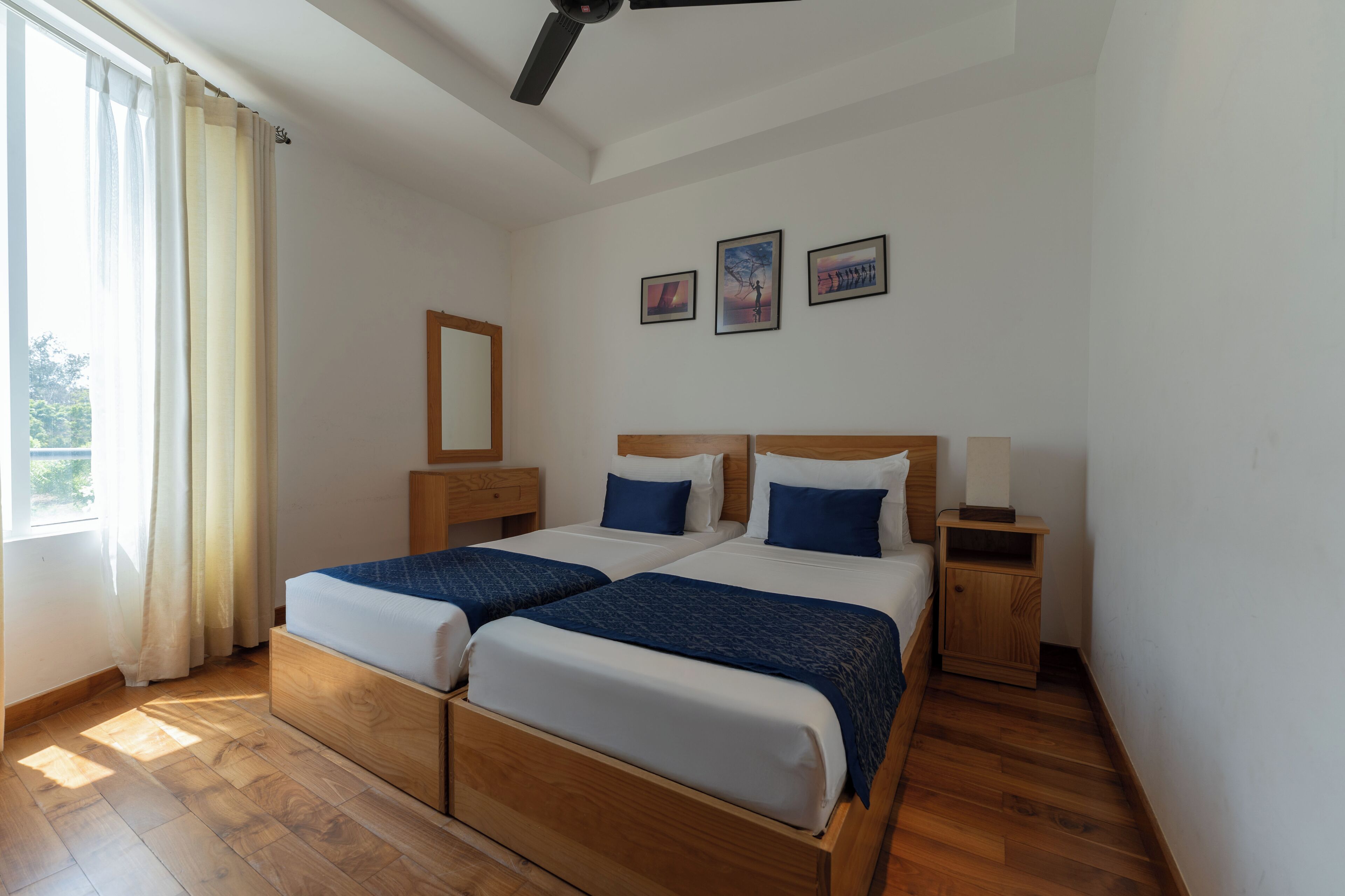 A bedroom with two beds, wooden flooring, a ceiling fan, and three framed pictures on the wall.