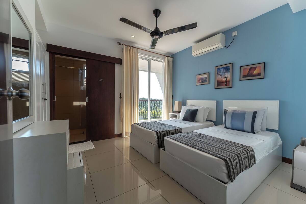 A hotel room with two beds, a ceiling fan, a large window with curtains, and artwork on the wall.