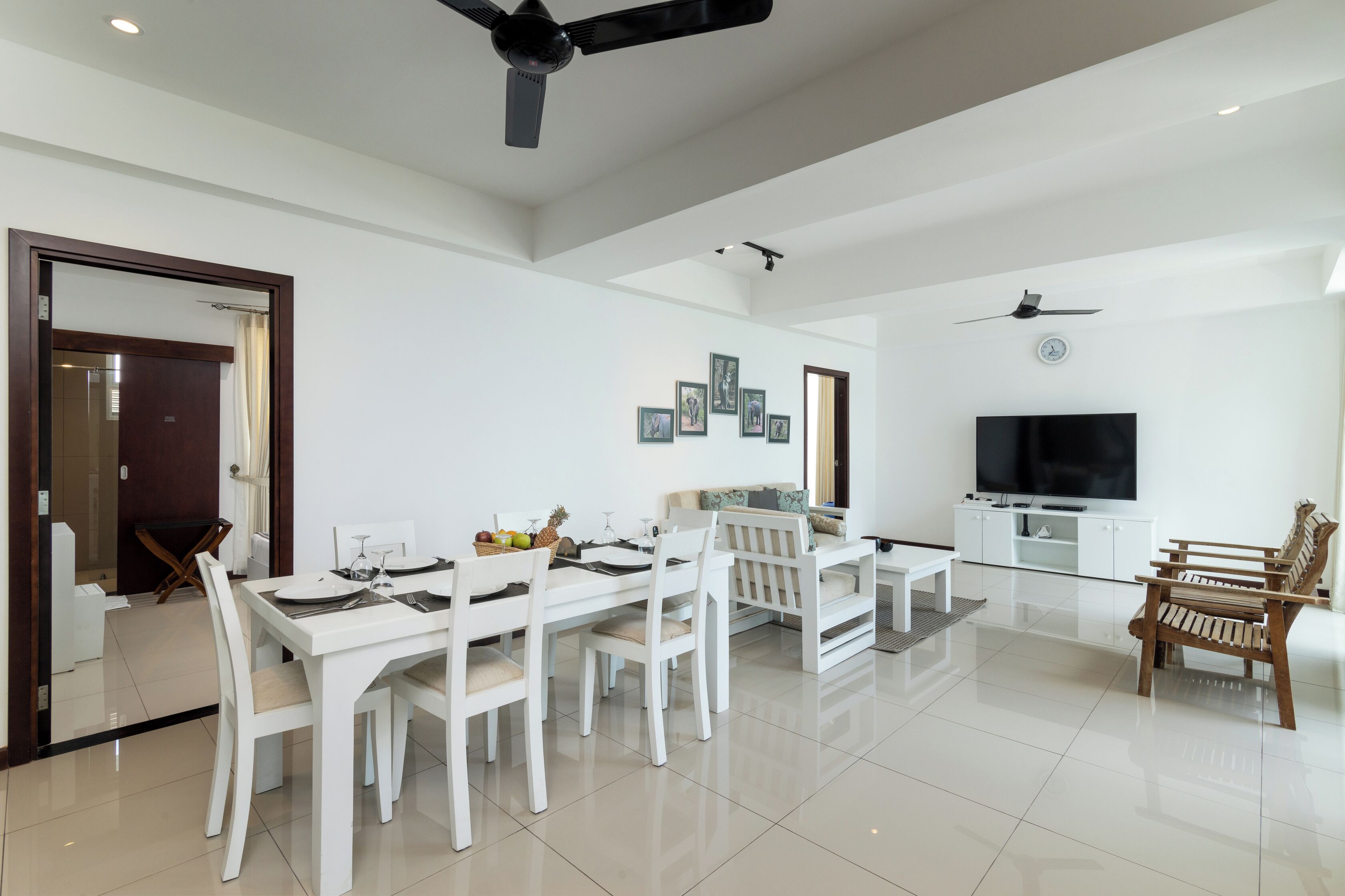 A modern living room with a dining area, white furniture, a flat-screen TV, and ceiling fans.