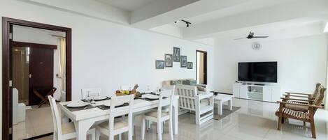 A modern living room with a dining area, white furniture, a flat-screen TV, and ceiling fans.