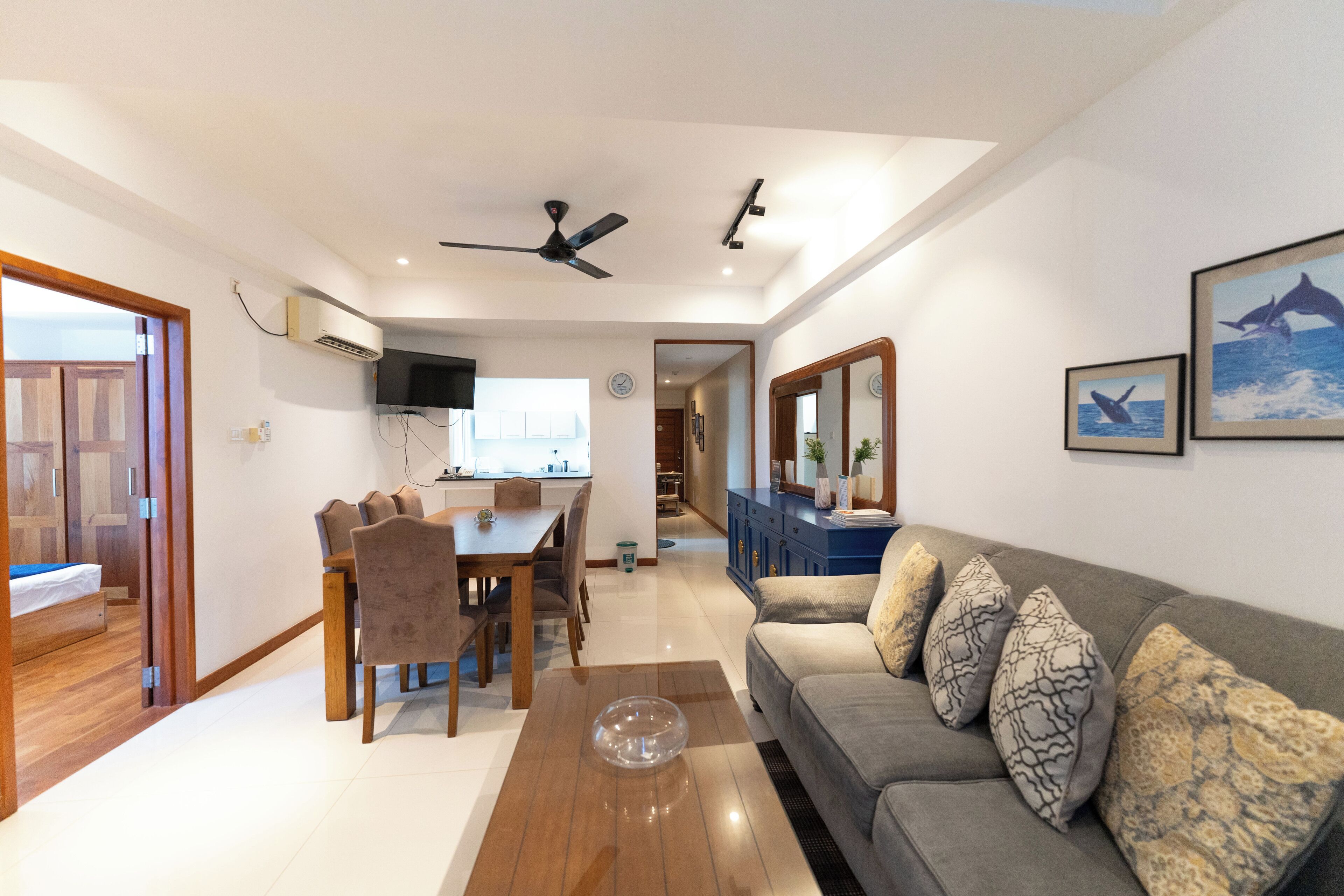 Un'ampia zona giorno con divano, tavolo da pranzo e sedie. Sono presenti un ventilatore a soffitto, una TV e un grande specchio a parete.