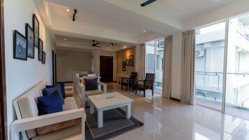 A spacious living area with a sofa, coffee table, and chairs. There is a balcony with a view of the outside.