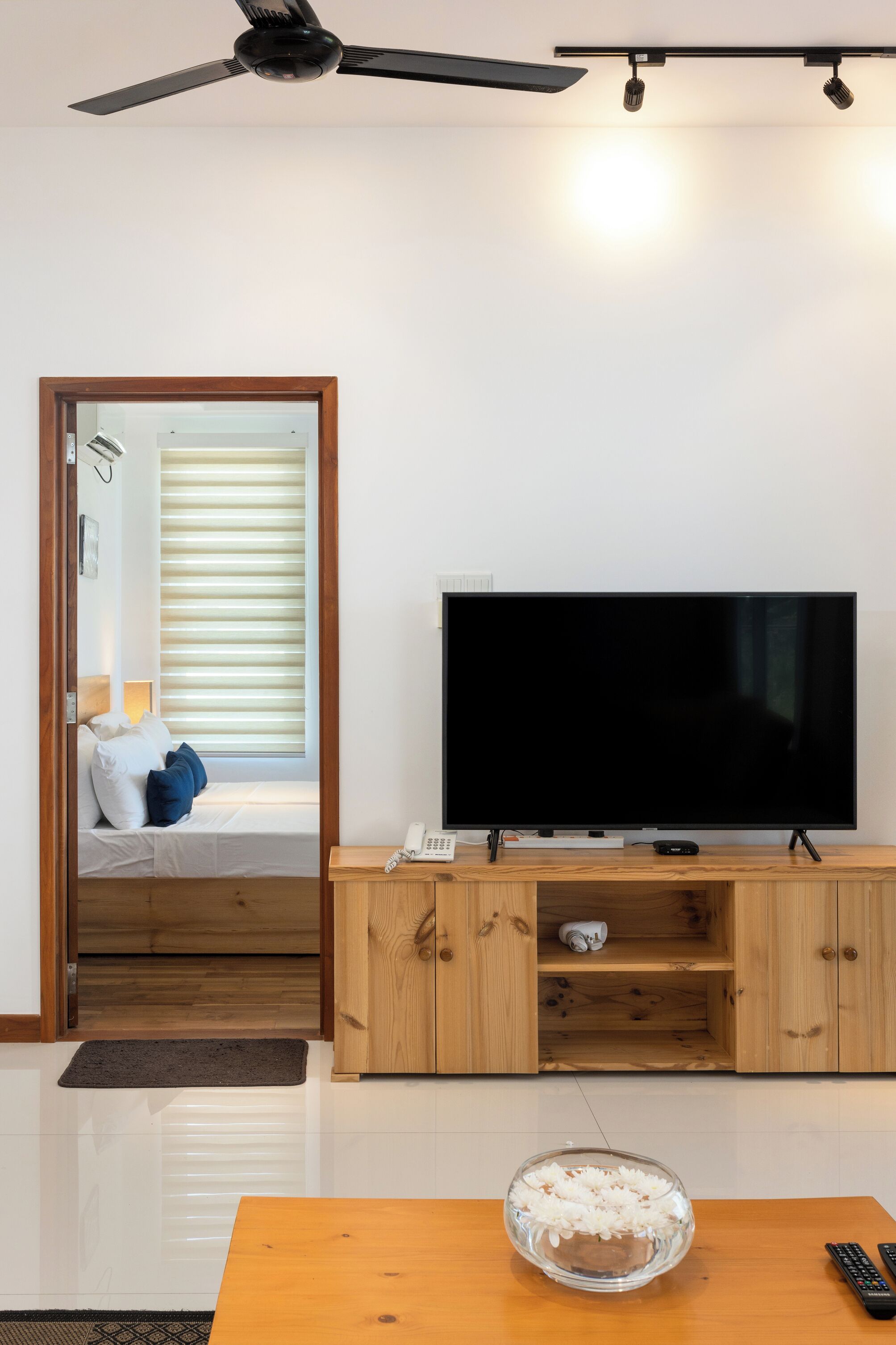 Een moderne woonkamer met een flatscreen televisie op een houten standaard, een kleine tafel met een schaal rijst, en een doorgang naar een slaapkamer met een bed en een raam met zonwering.