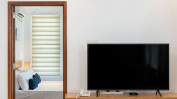 Een moderne woonkamer met een flatscreen televisie op een houten standaard, een kleine tafel met een schaal rijst, en een doorgang naar een slaapkamer met een bed en een raam met zonwering.