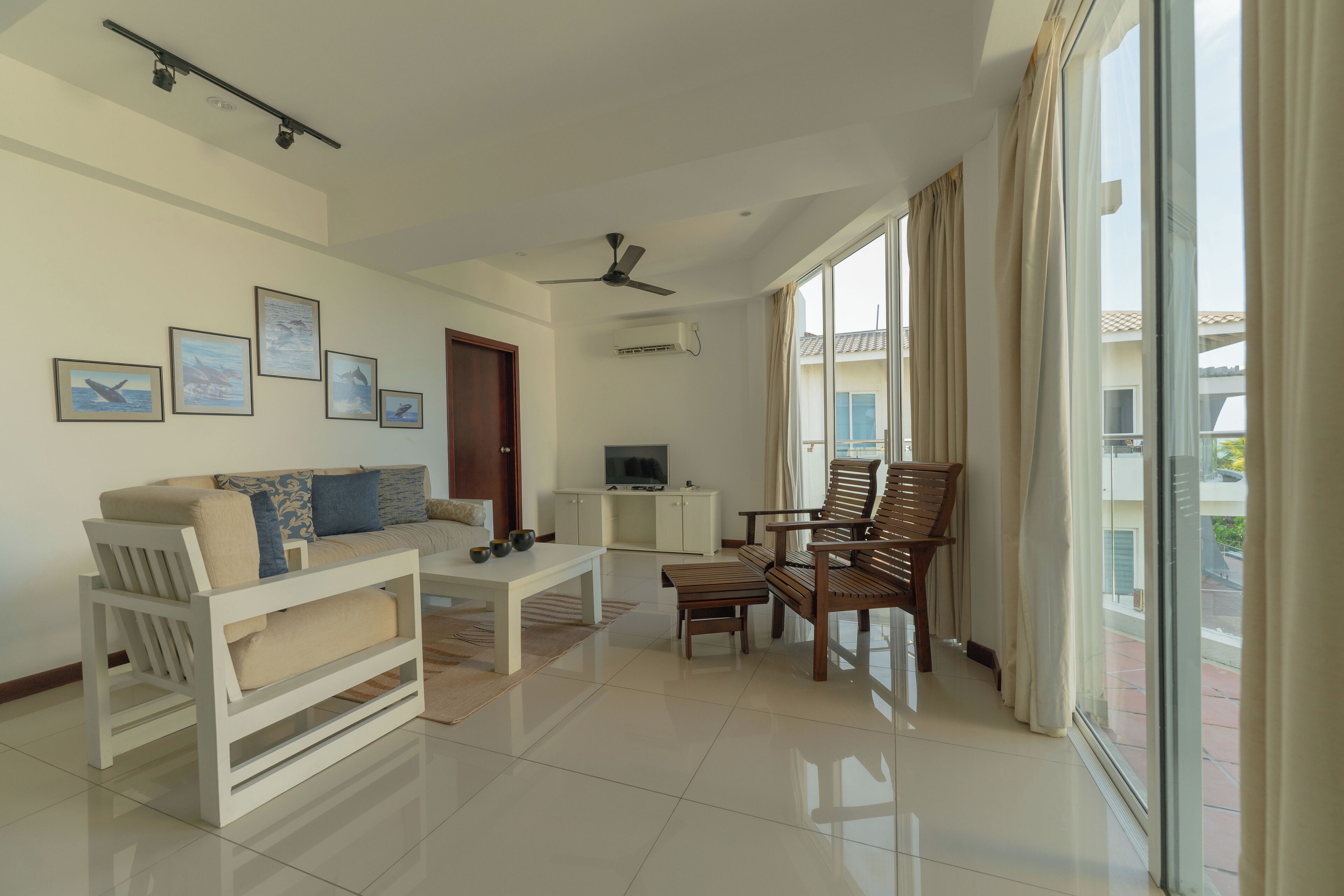 A spacious living room with a sofa, coffee table, and wooden chairs. There are framed pictures on the wall, a ceiling fan, and a large window with curtains.