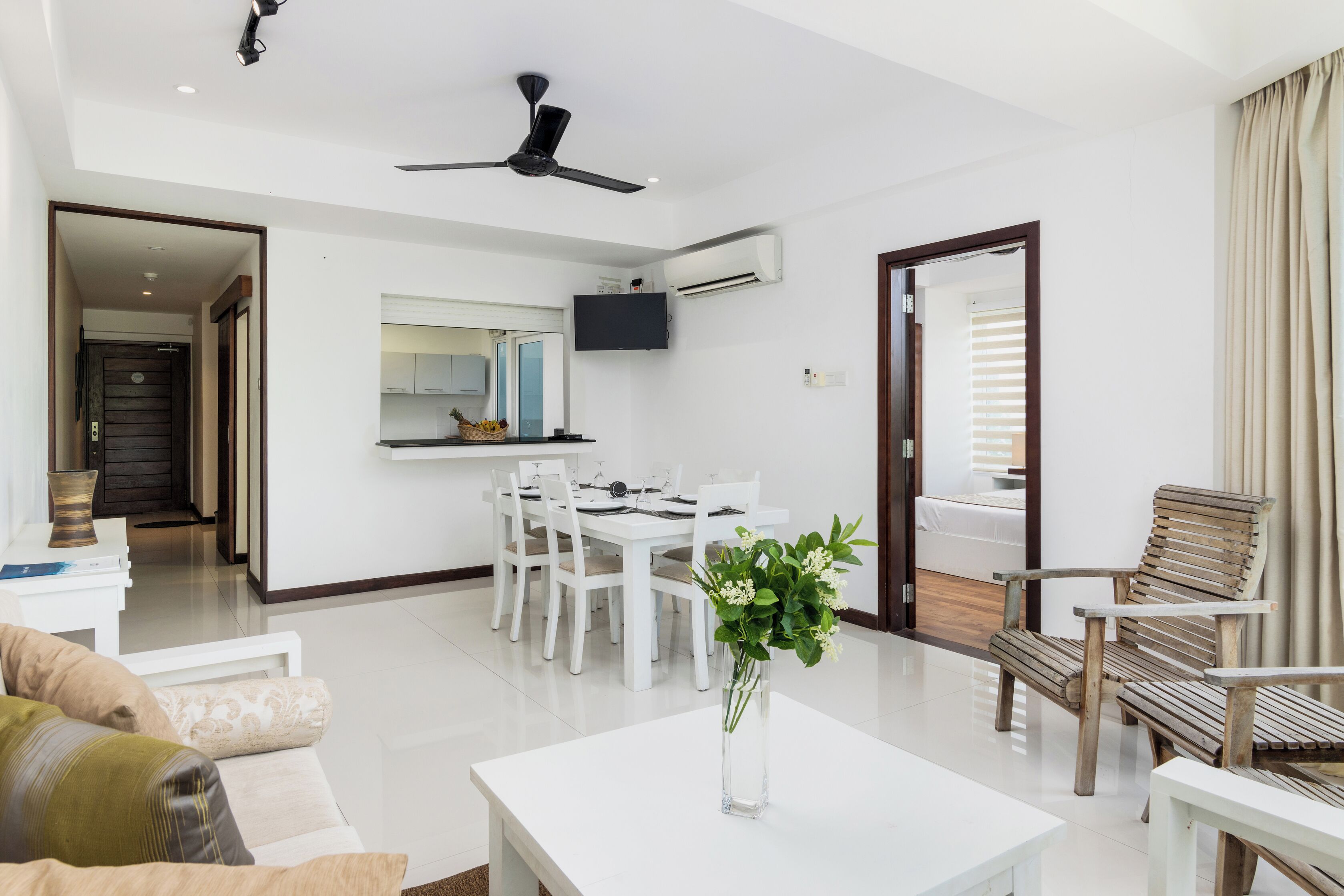 Uma sala de estar moderna com um sofá branco, uma mesa de centro e uma cadeira de madeira. Há uma área de jantar com uma mesa e cadeiras brancas, e uma cozinha visível ao fundo.