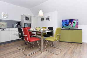 Uma cozinha moderna com armários brancos, micro-ondas e máquina de café. Há uma mesa de jantar com cadeiras coloridas e uma televisão exibindo um desenho animado.