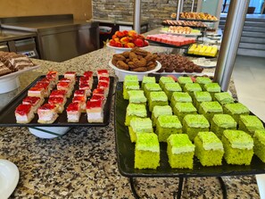 A buffet with various desserts, including red-topped and green-topped cakes, arranged on a granite countertop. - Nessebar Beach Hotel (Nessebar)