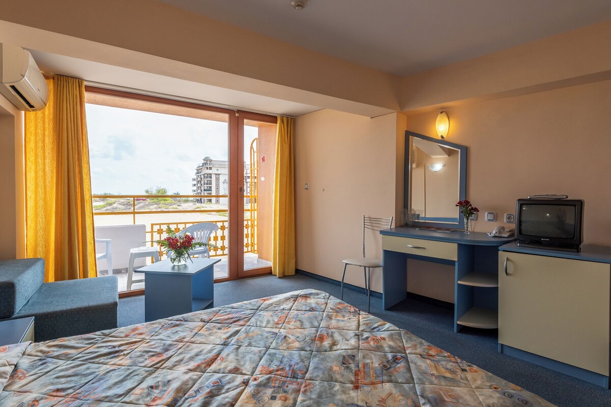 A hotel room with a bed, a desk, a chair, a TV, and a balcony with yellow curtains.