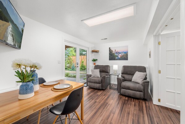 A modern living room with a wooden dining table, two grey armchairs, a grey sofa, a TV mounted on the wall, and a cityscape painting.
