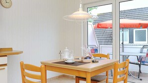 A dining area with a wooden table and chairs, a white ceiling light, and a view of the outside through a glass door. - Apartment "top right" - House Pamir (Neuharlingersiel)