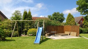 A backyard with a wooden swing set, a blue slide, a wooden bench, and a small table. - Apartment "top right" - House Pamir (Neuharlingersiel)