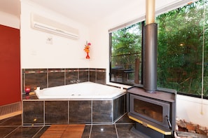 A modern bathroom with a large bathtub, a wood-burning stove, and a window with a view of greenery.