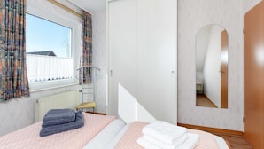 Ein Schlafzimmer mit Bett, einem Fenster mit Vorhängen, einem Kleiderschrank, einem Spiegel und einem Heizkörper.