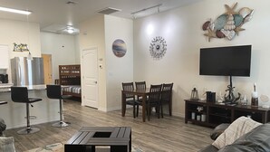 A modern living room with a leather sofa, a dining area with wooden chairs, a kitchen with a stainless steel refrigerator, and a flat-screen TV mounted on the wall. - 1b/1b for 6 max4adults! Heated pools! Steps from the BEACH! Netflix! Playground! (Corpus Christi)