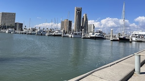 A marina with multiple boats docked, a clear sky, and modern buildings in the background. - 1b/1b for 6 max4adults! Heated pools! Steps from the BEACH! Netflix! Playground! (Corpus Christi)