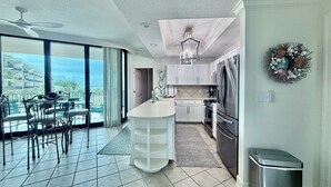 Een moderne keuken met een groot eiland, een zithoek met stoelen, uitzicht door grote ramen en een plafondventilator.