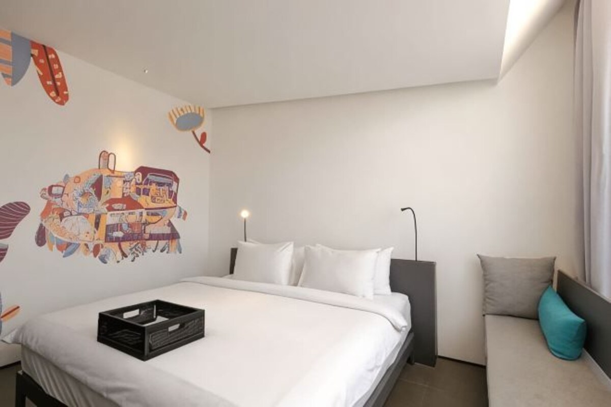 A modern hotel room with a bed, a sofa, a wall art piece, and a window with curtains.