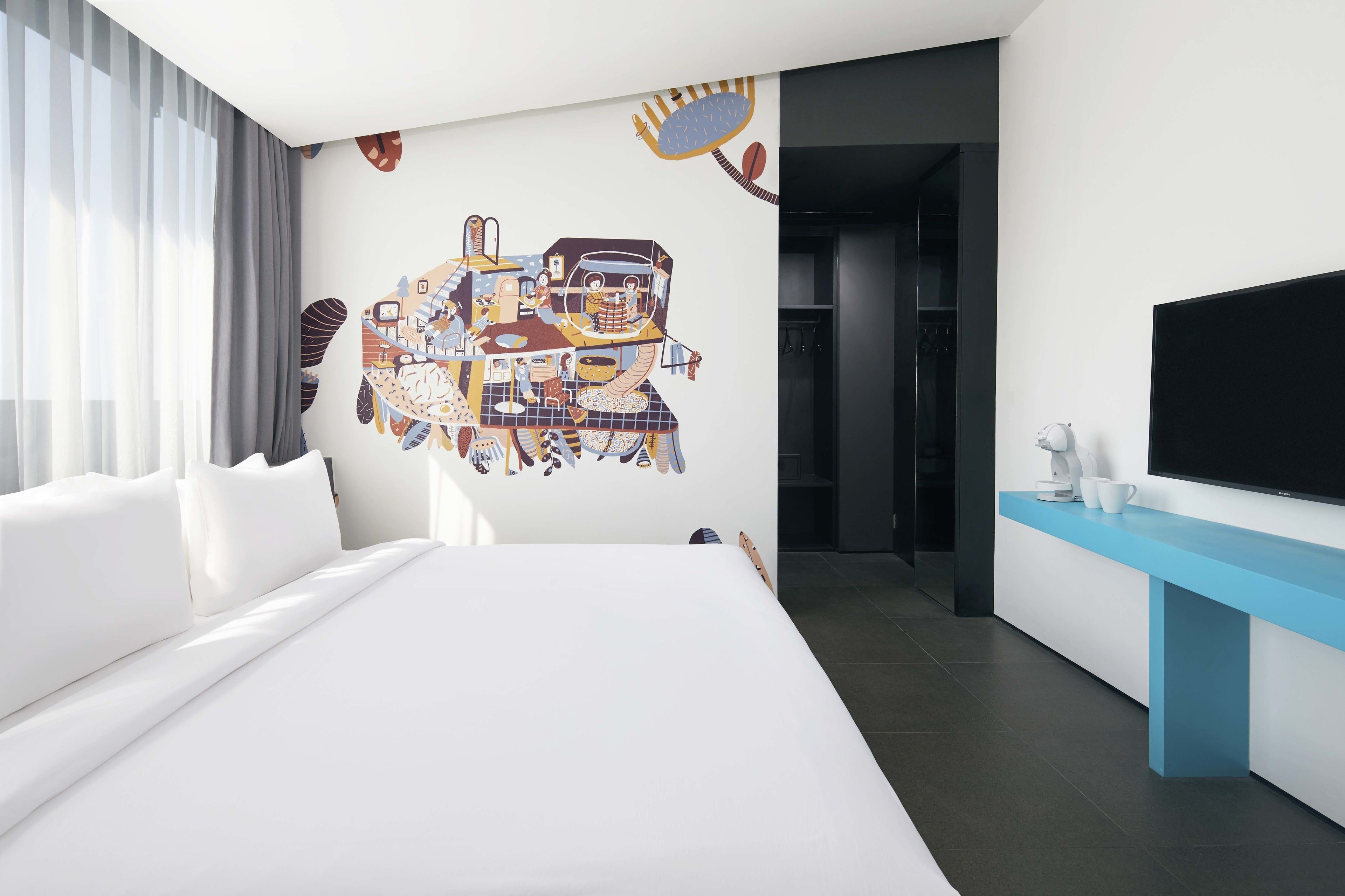 Une chambre d’hôtel moderne avec un grand lit, un téléviseur à écran plat, une console bleue et une fresque murale aux couleurs vives.