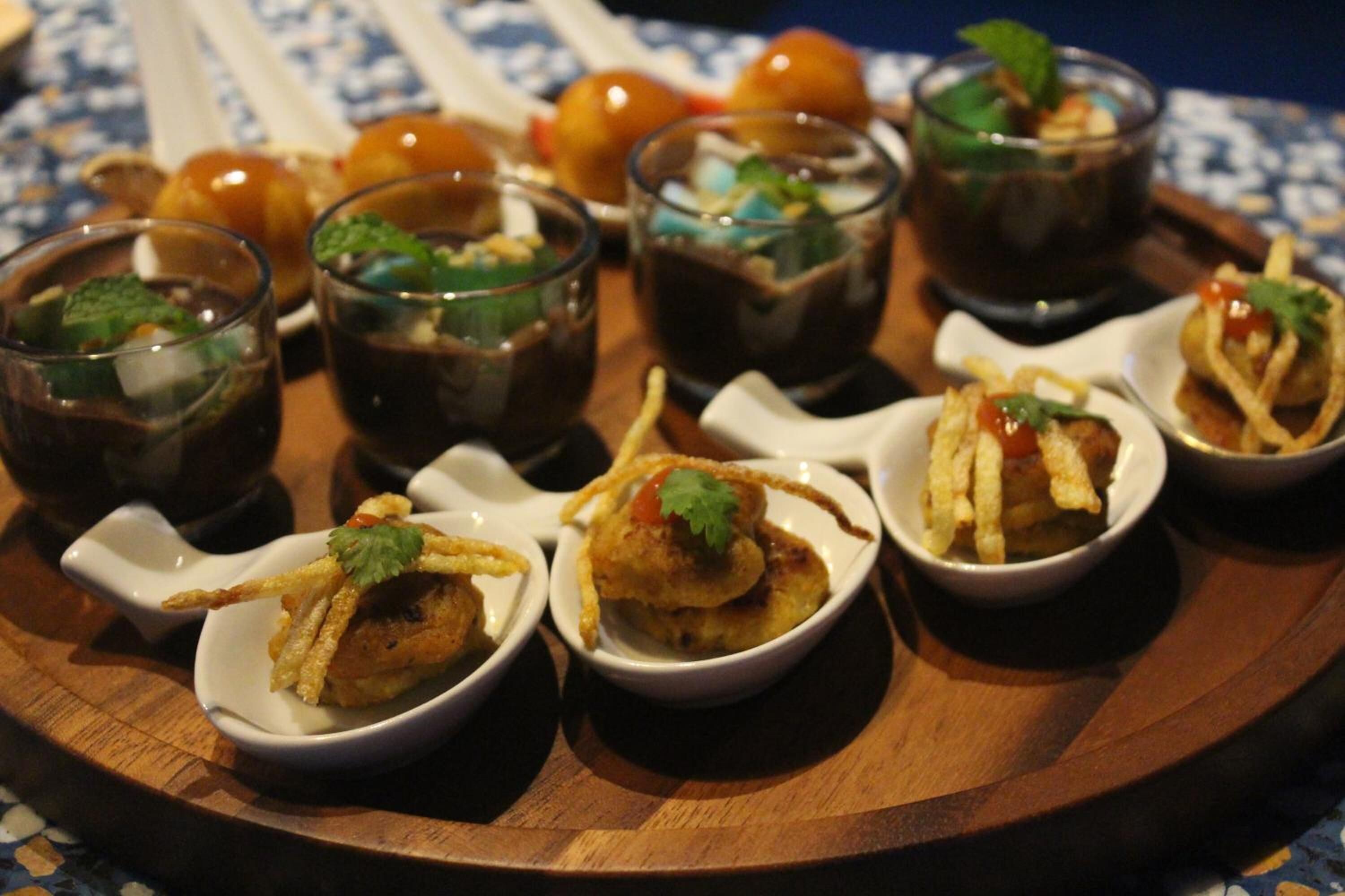 Un plateau contenant de petits bols remplis de nourriture et de boissons, notamment un plat agrémenté d’une garniture et un verre contenant un liquide sombre.