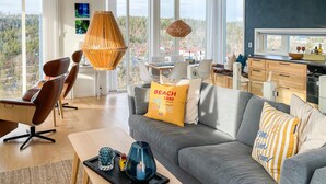 Un salon moderne avec un canapé gris, un fauteuil en cuir marron et un coin repas comprenant une table et des chaises. Une grande fenêtre offre une vue sur un paysage boisé.