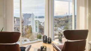 Une pièce moderne avec de grandes fenêtres donnant sur un paysage urbain et une étendue d’eau. On y trouve deux fauteuils en cuir et deux chandeliers noirs posés sur le rebord de la fenêtre.