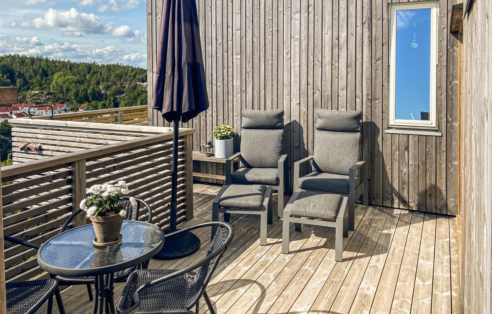 Una terraza de madera con una mesa, sillas, una planta en maceta y un ventanal con vistas a un área boscosa.