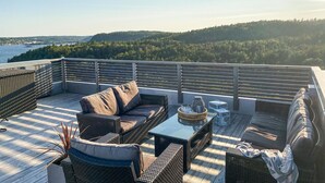 Une terrasse dotée de meubles en osier, d’une table en verre et offrant une vue sur un paysage boisé.