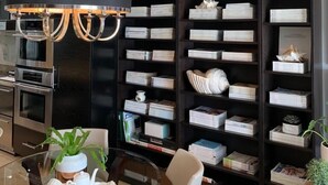 Ein modernes Wohnzimmer mit einer schwarzen Bücherwand, gefüllt mit Büchern und Dekoration, einem runden Glastisch mit Teekanne und Tassen sowie einer Kronleuchter.