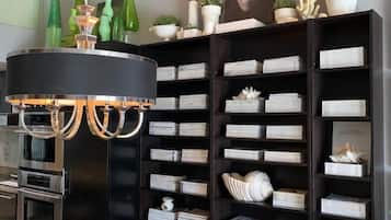 Una sala moderna con una estantería negra llena de libros y objetos decorativos, una mesa de centro redonda de vidrio con una tetera blanca y tazas, y una araña.