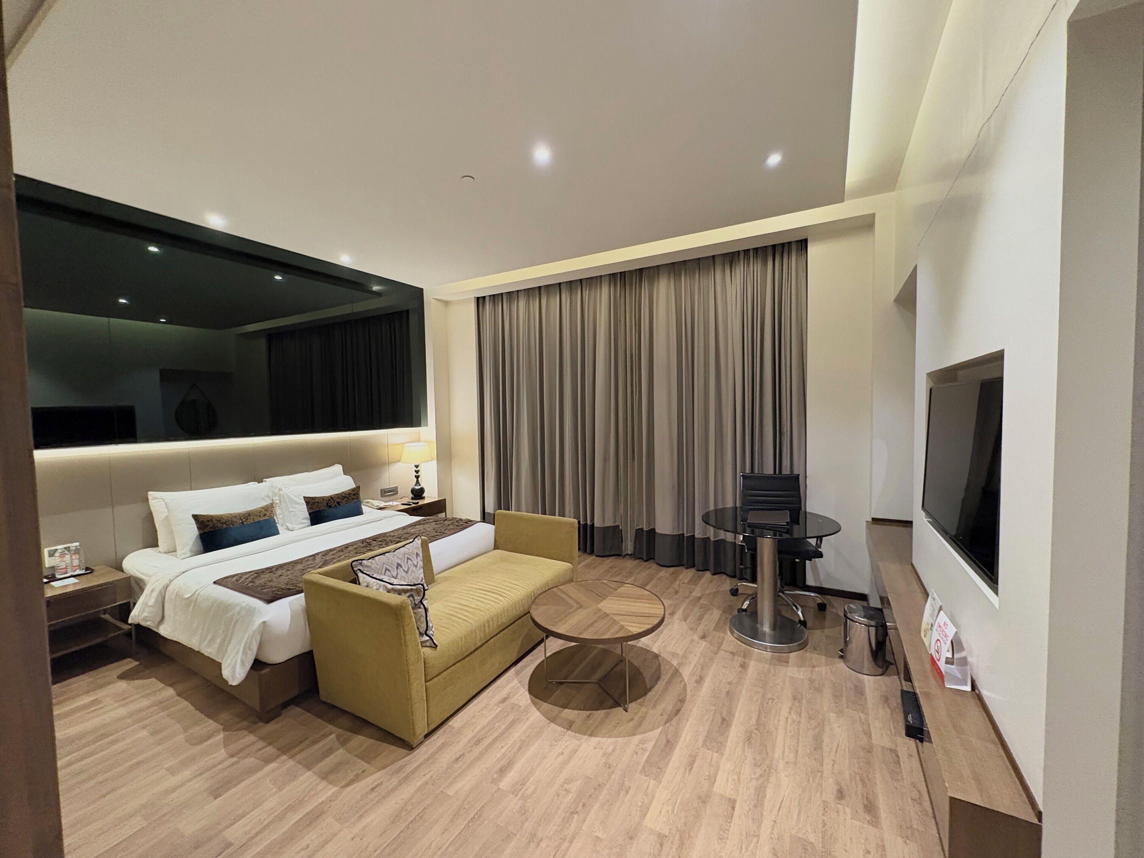Une chambre d’hôtel moderne équipée d’un lit, d’un canapé, d’une table basse, d’une télévision et d’une grande fenêtre avec des rideaux.