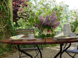A garden table with a vase of flowers, a teapot, and cups, surrounded by greenery and a bench. - Apartment Schleiwiese - Apartments Hartmann (Rabenkirchen-Faulück)