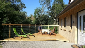 Une terrasse en bois avec une table et des chaises, entourée d’une clôture en métal, et un bâtiment en briques en arrière-plan.