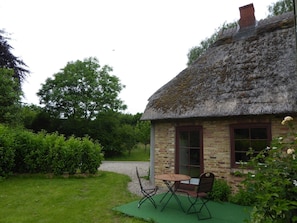 A thatched-roof cottage with a brick exterior, a small patio with a table and chairs, and a garden with bushes and trees.