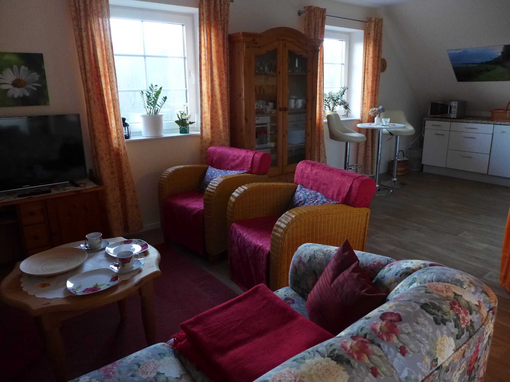 A living room with a television, wicker furniture, a dining table with plates, and a kitchen area with cabinets and a microwave.