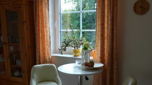 A room with a window, two chairs, a table with a plant and fruit, and a clock on the wall.