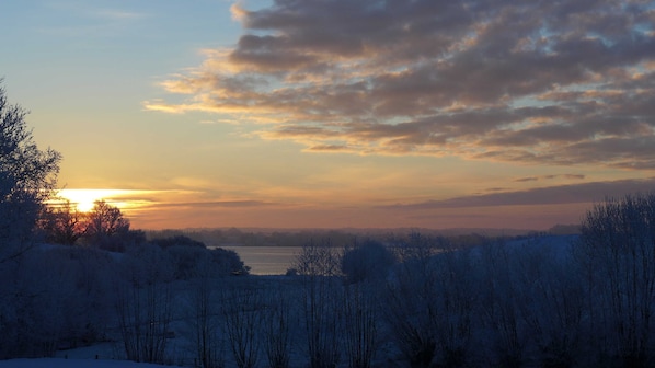 A winter landscape with a sunset, snow-covered trees, and a body of water. - Apartment Schleiwiese - Apartments Hartmann (Rabenkirchen-Faulück)