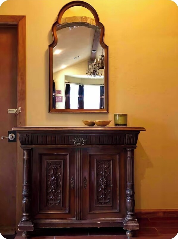 Una consola de madera con un espejo encima, una puerta a la izquierda y un candelabro al fondo.