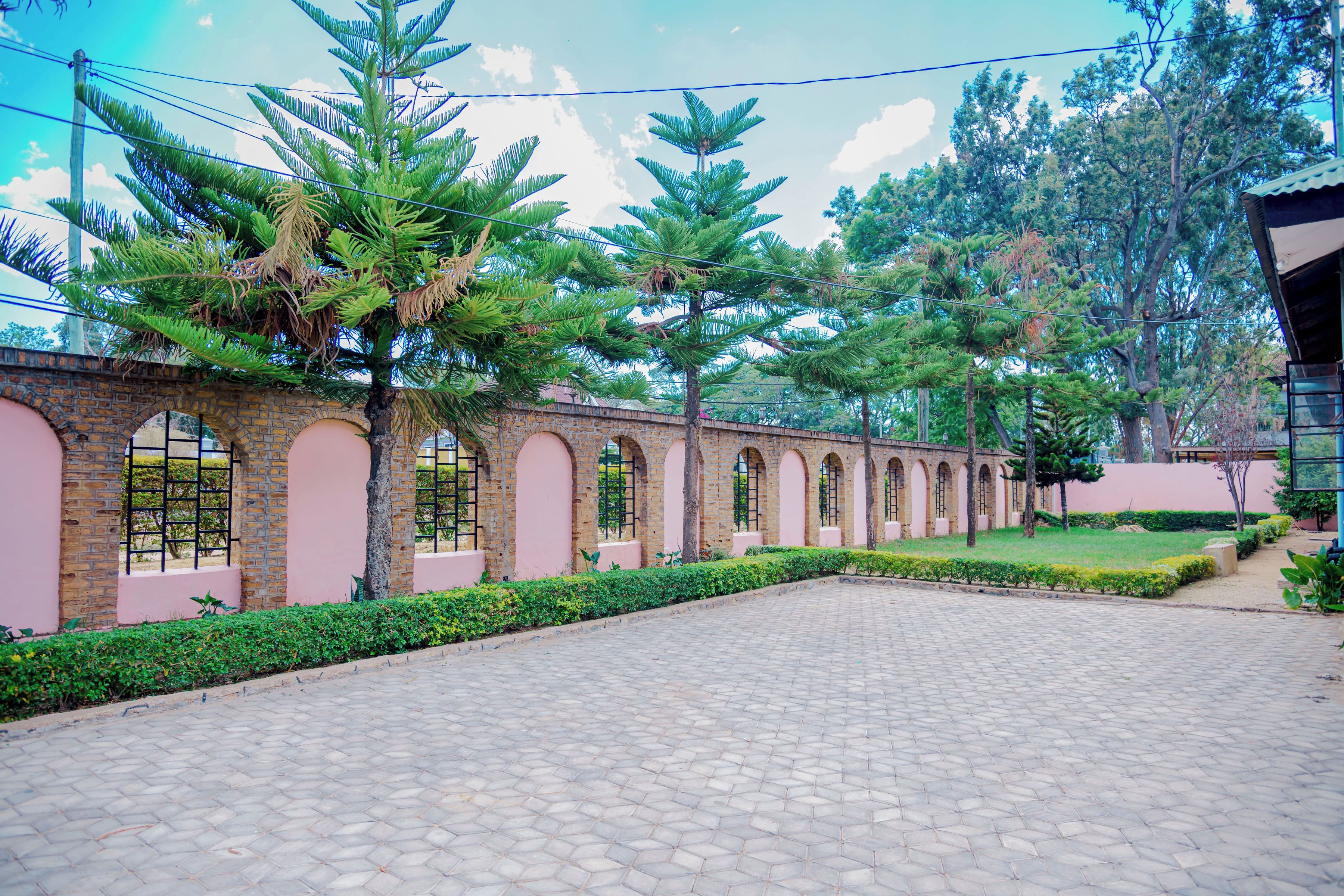 A paved courtyard with a brick wall, arched windows, and a row of trees.