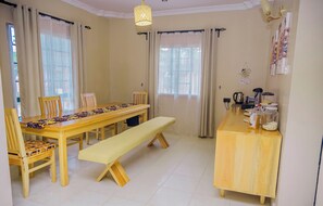 Un espace repas avec une longue table en bois, des bancs et des chaises. On y trouve un petit comptoir en bois équipé d’appareils de cuisine et d’une machine à café.