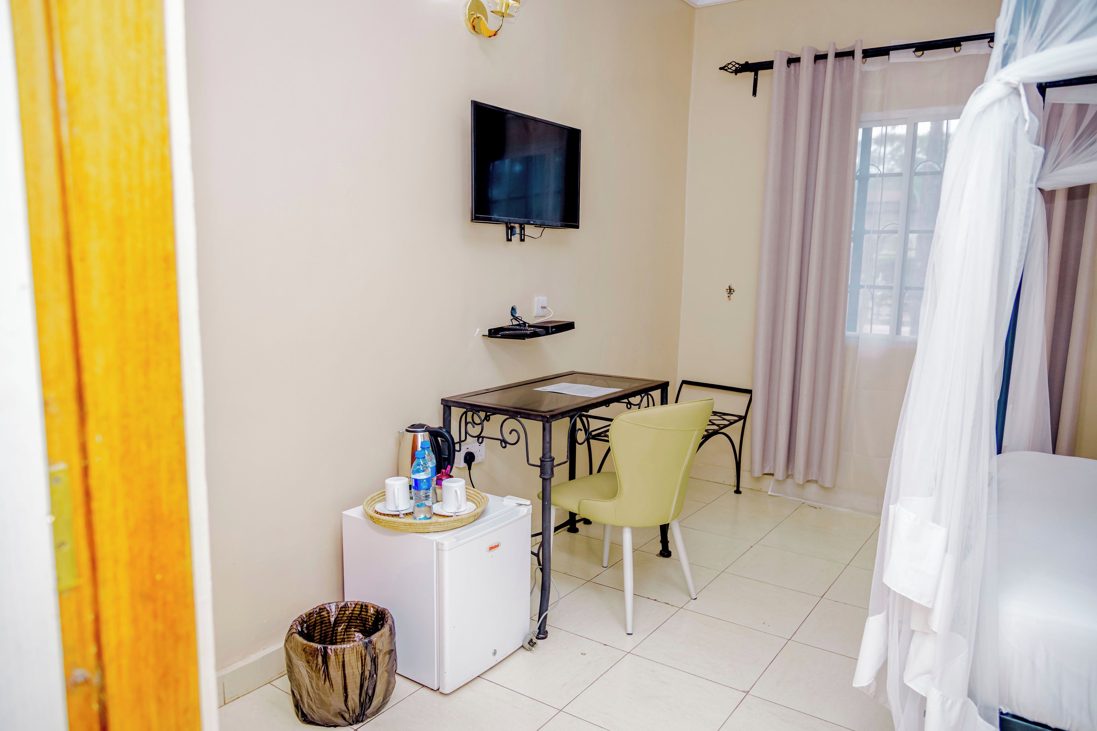 A small, clean room with a bed, a television mounted on the wall, a table with chairs, and a trash bin.