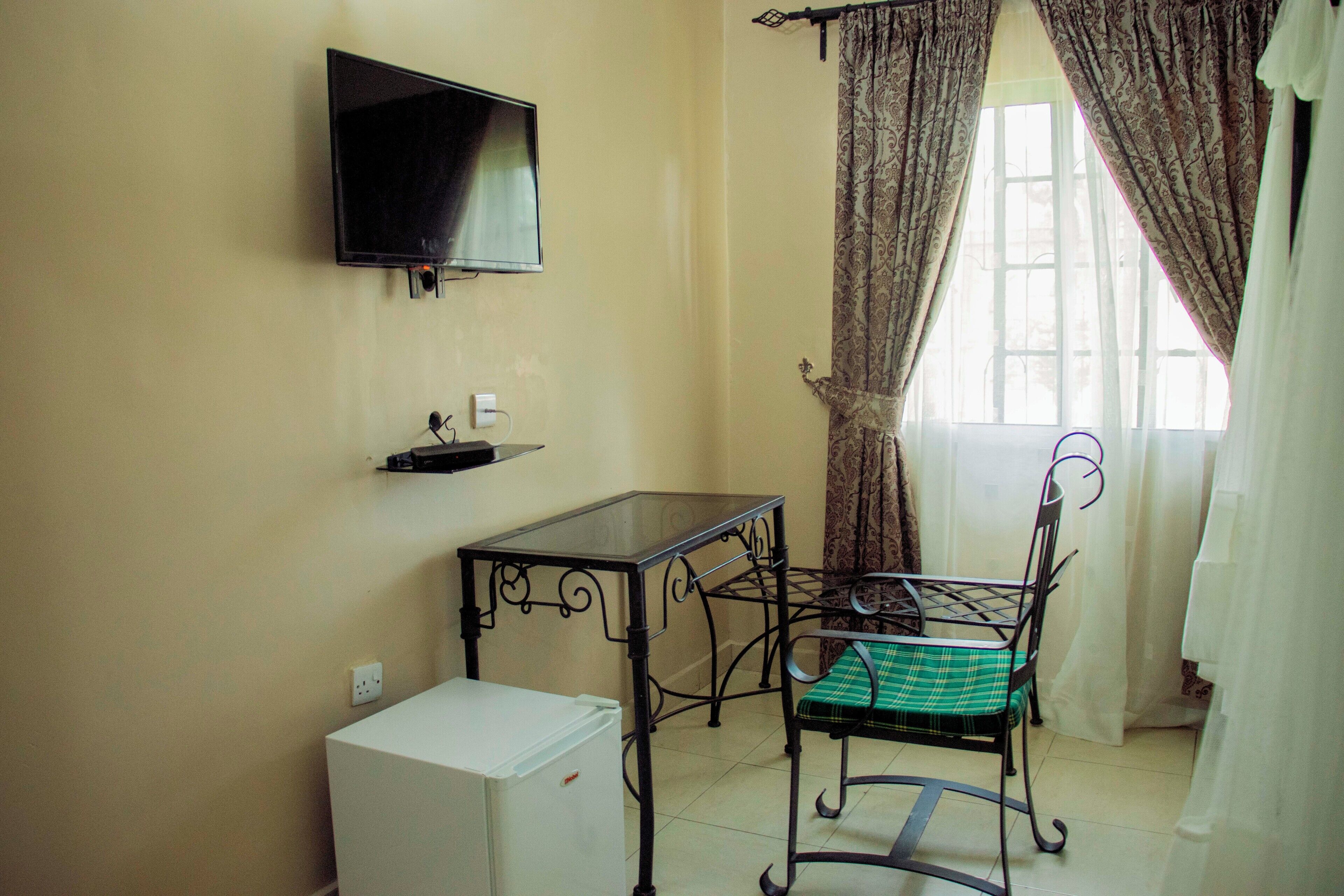Una habitación pequeña con una televisión montada en la pared, una mesa con una silla, un refrigerador pequeño y una ventana con cortinas.
