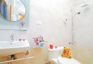 A bathroom with a white sink, mirror, toilet, and shower. There is a teddy bear on the toilet seat and a pink flower vase on the sink. - Happiness Garden (Ji'an)