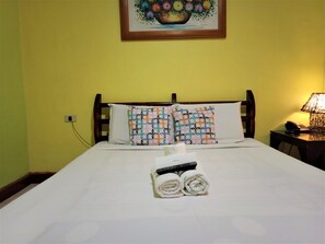 Una cama bien hecha con sábanas blancas, dos almohadas estampadas, una toalla enrollada y un control remoto.