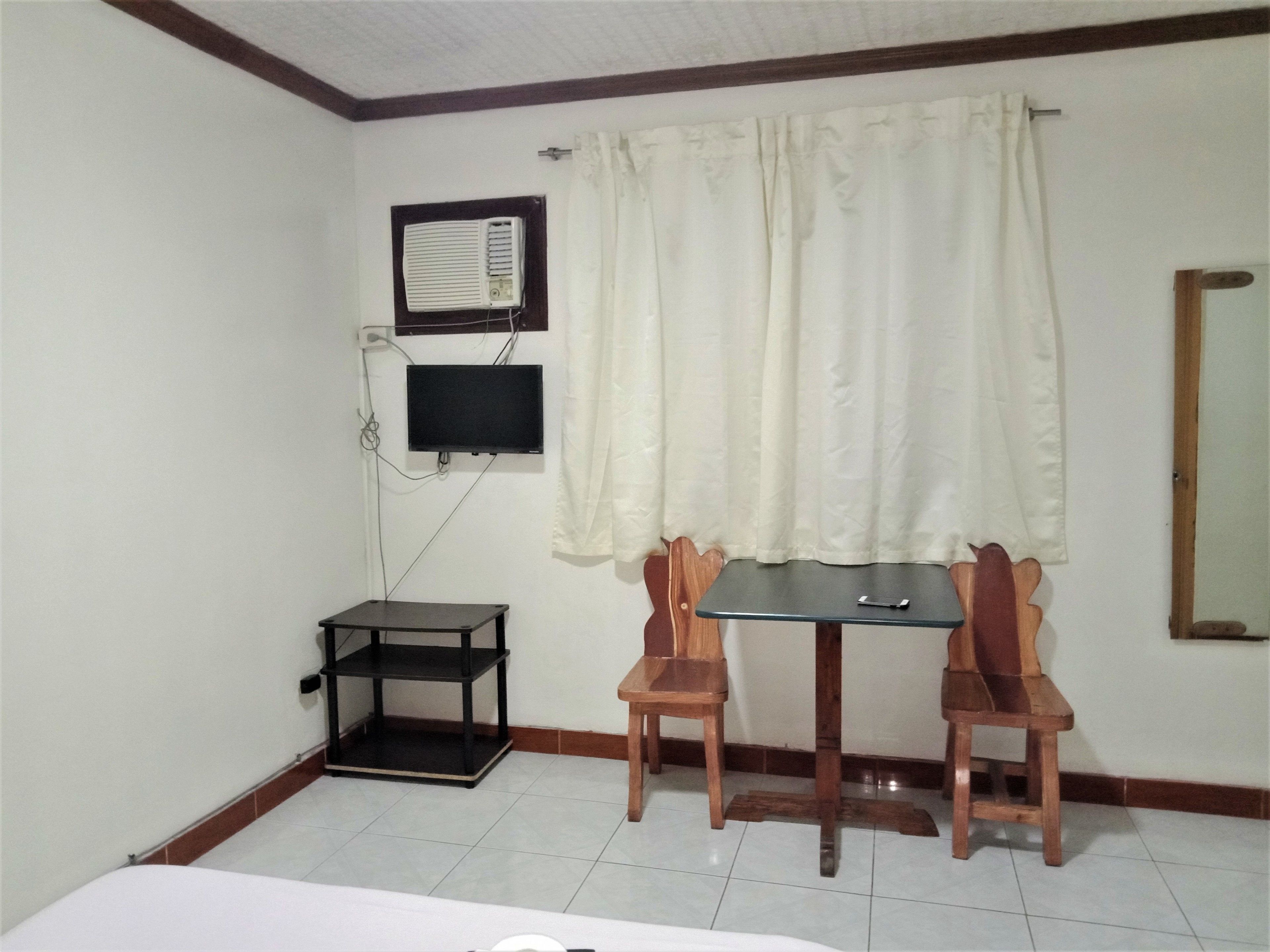 A small room with a bed, a TV mounted on the wall, a table with two chairs, and a window with curtains.