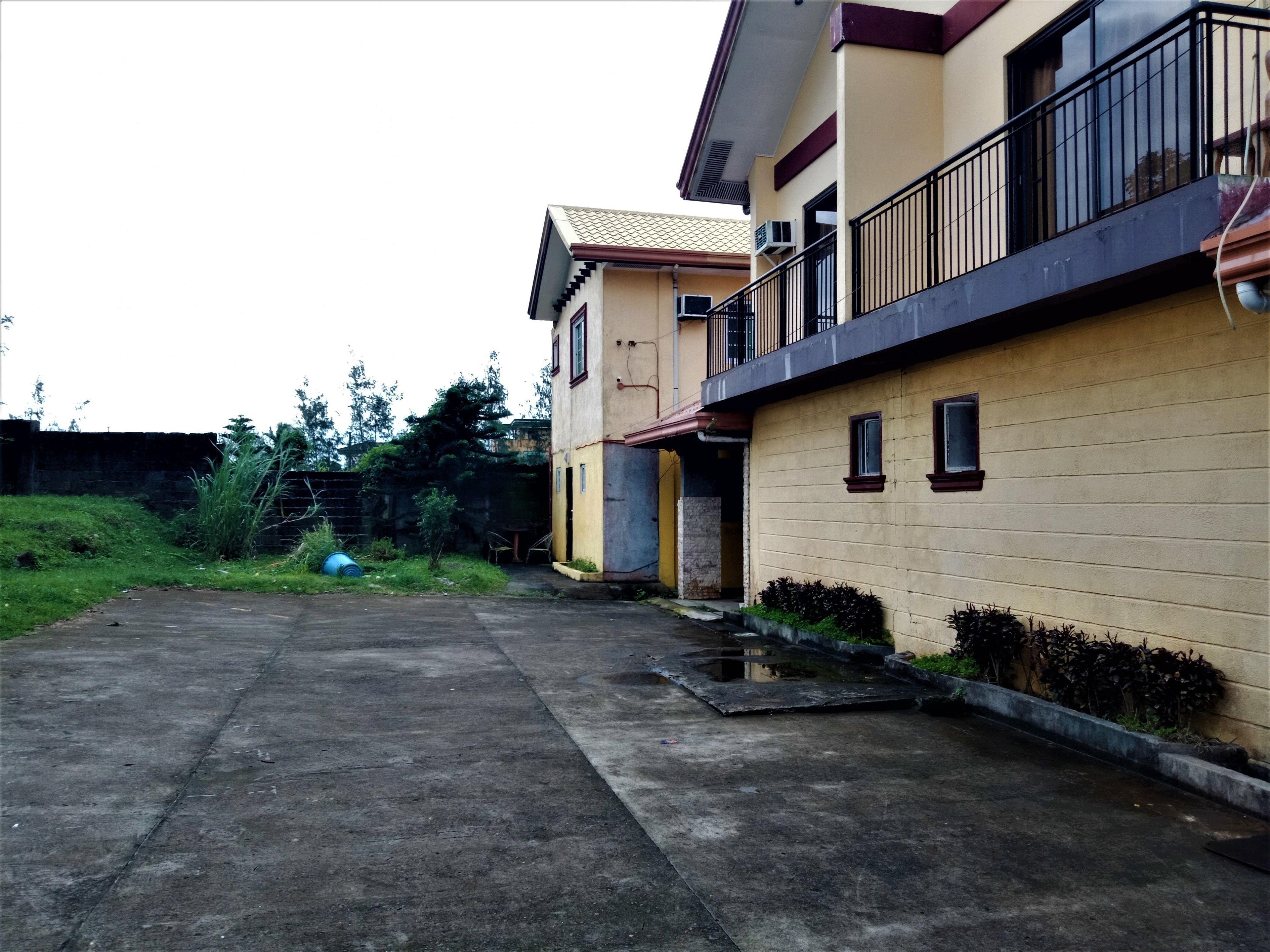 A two-story residential building with a balcony, a concrete driveway, and a small garden area.