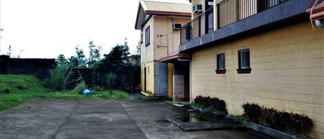 A two-story residential building with a balcony, a concrete driveway, and a small garden area.