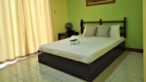 A hotel room with a bed, a nightstand, a lamp, a painting on the wall, and a window with curtains. - La Gracia Apartelle Tagaytay (Tagaytay)
