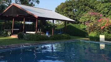 A pool with a covered pavilion, surrounded by lush greenery and flowering plants.