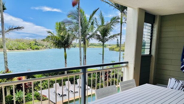 A balcony with a table and chairs, overlooking a body of water and palm trees. - Kingscliff Waterfront - large pool, direct creek access & 5 mins walk to beach (Kingscliff)